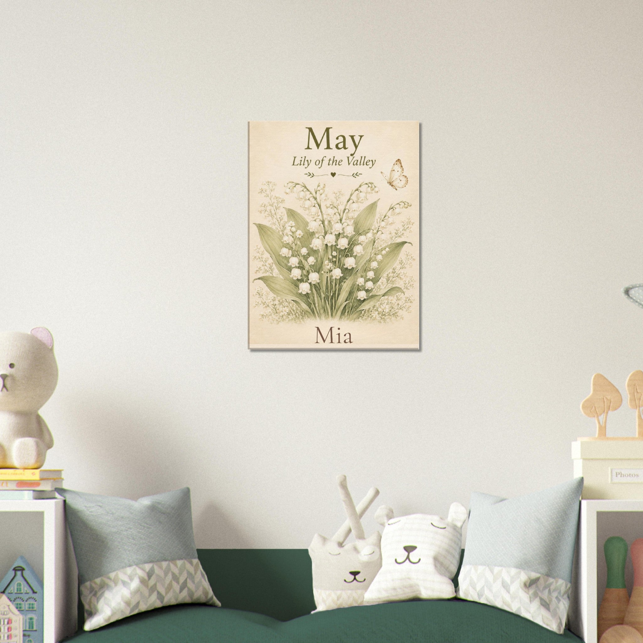 A personalized May floral wall art print hanging above a green sofa in a styled children's room. The print features lily of the valley flowers with the text 'May - Lily of the valley - Mia' on a cream background.