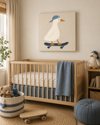 A styled nursery room featuring a light wood crib with a blue quilt and mattress, a blue striped storage basket with teddy bears, a blue pouf, and a canvas wall art print of a white duck on a skateboard wearing a blue cap.