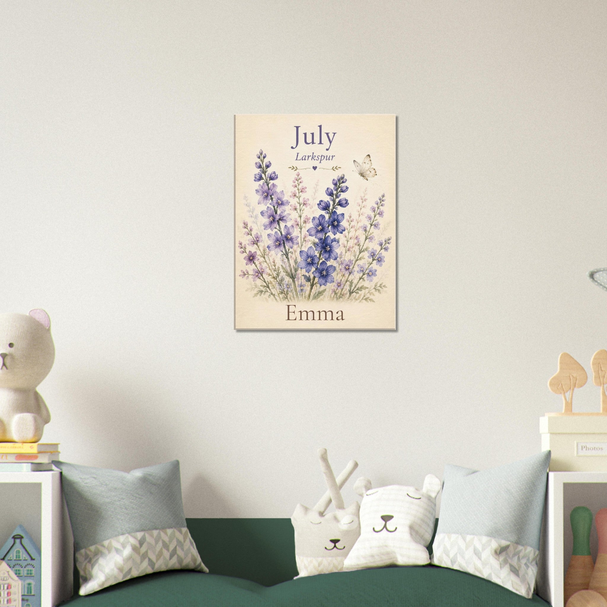 A personalized July flower wall art print featuring lavender and delphinium illustrations with the name 'Emma' at the bottom, displayed on a light wall above a green sofa with decorative pillows.