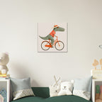 A square wall art print hanging above a green sofa in a styled room. The print shows a cartoon dinosaur riding an orange bicycle against a pale pink background.
