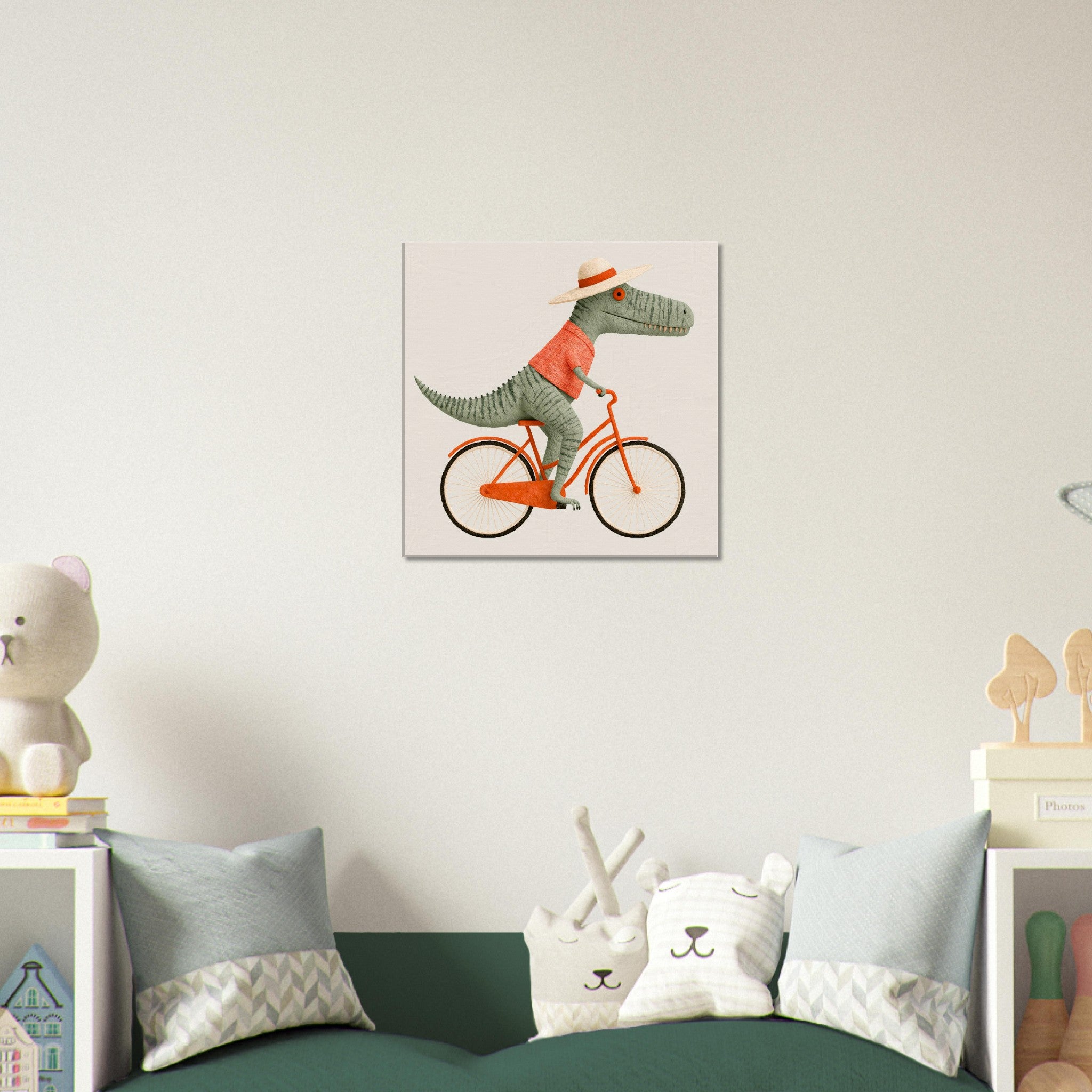 A square wall art print hanging above a green sofa in a styled room. The print shows a cartoon dinosaur riding an orange bicycle against a pale pink background.