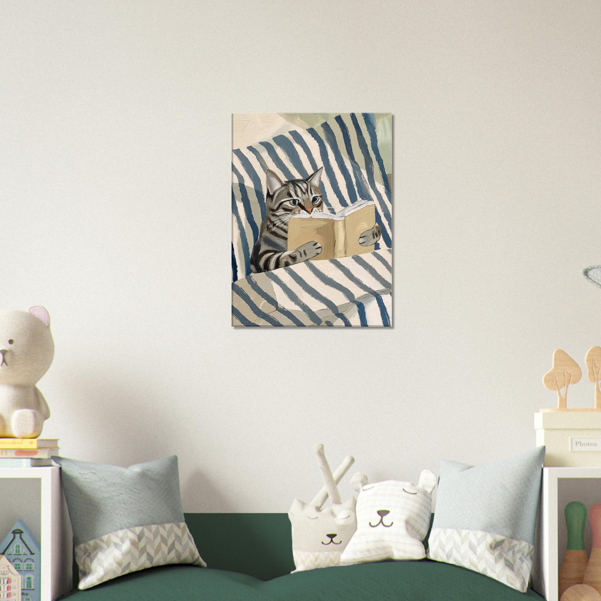 A framed wall art print showing a tabby cat reading a book on blue-and-white striped bedding, displayed above a sofa in a styled room with pillows and small decor items.