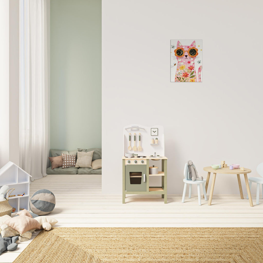 A styled children's playroom with a floral cat wall art print hanging on a white wall above a small wooden play kitchen, chair, and table. The room features light wood floors, a woven rug, and a soft green accent wall in the background.